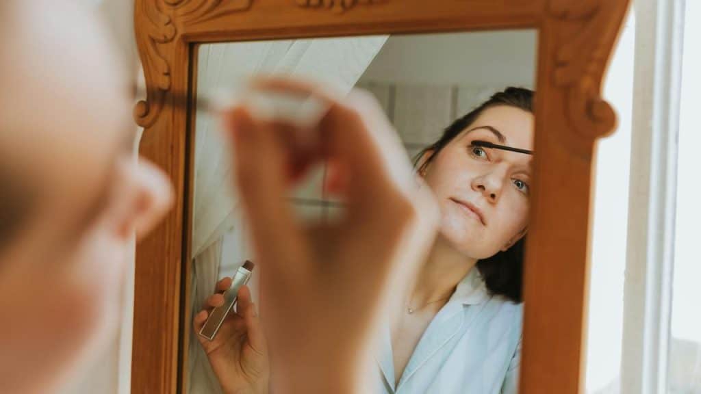 A woman applying mascara while looking into a mirror.