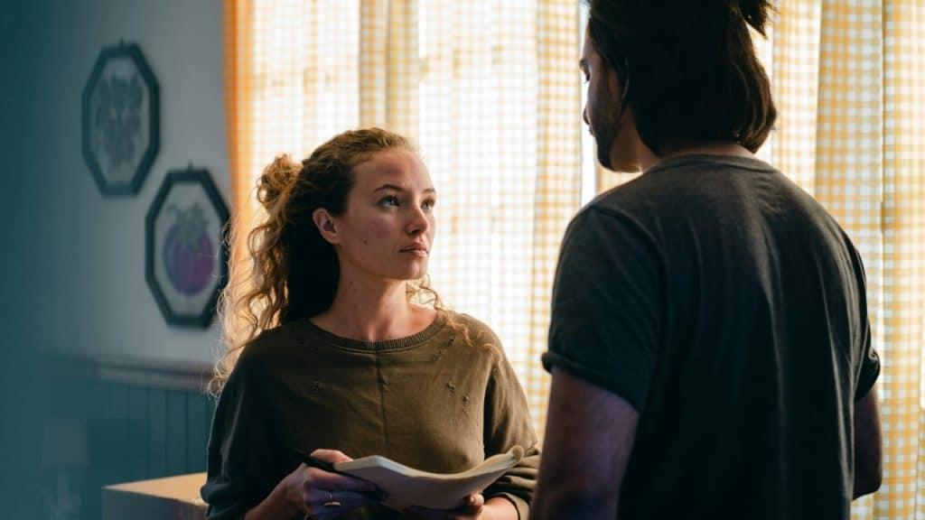 A woman with curly hair holding a notepad looks upward at a man standing before her.