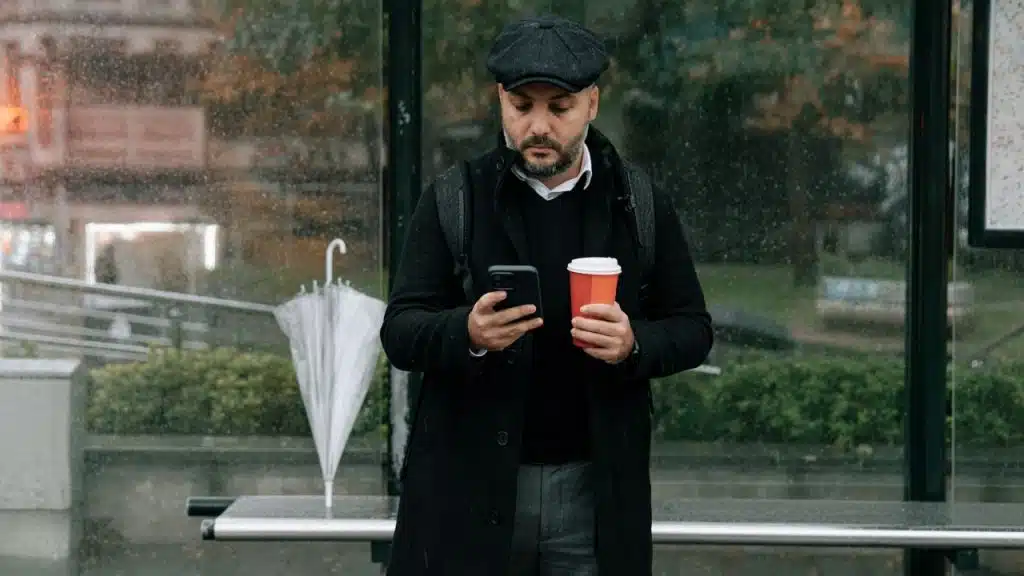 A man in a black coat and flat cap holds a cup while looking at a smartphone.