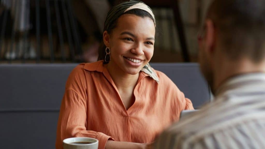 A woman in an orange shirt smiles while looking at a man across a table.