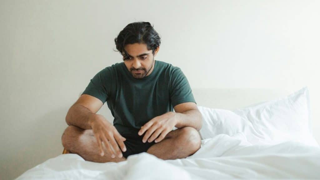 A man in a green shirt sits cross-legged on a white bed looking downward.