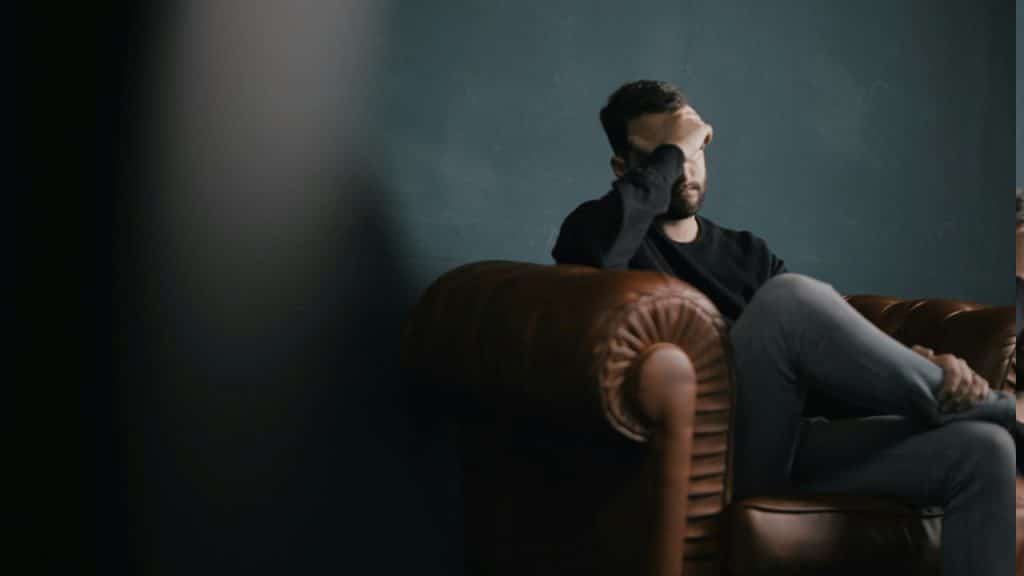 A depressed man holding his head with his hand while sitting on a sofa.