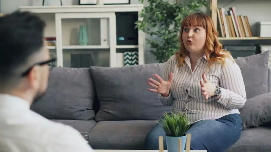 A woman with red hair gestures with open hands while sitting on a grey couch.