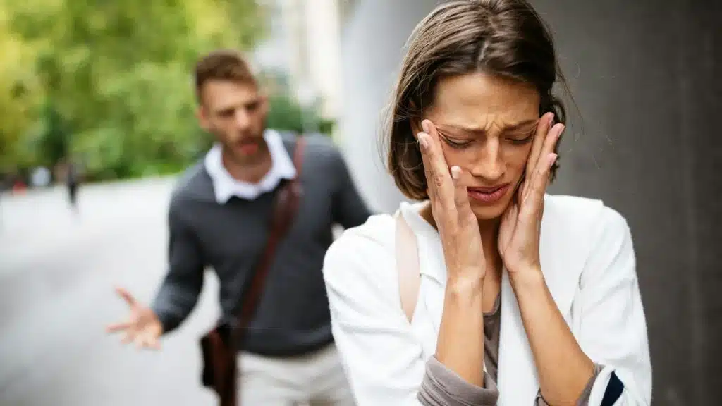 A woman holds a paper and speaks to a man who is resting his head in his hand.