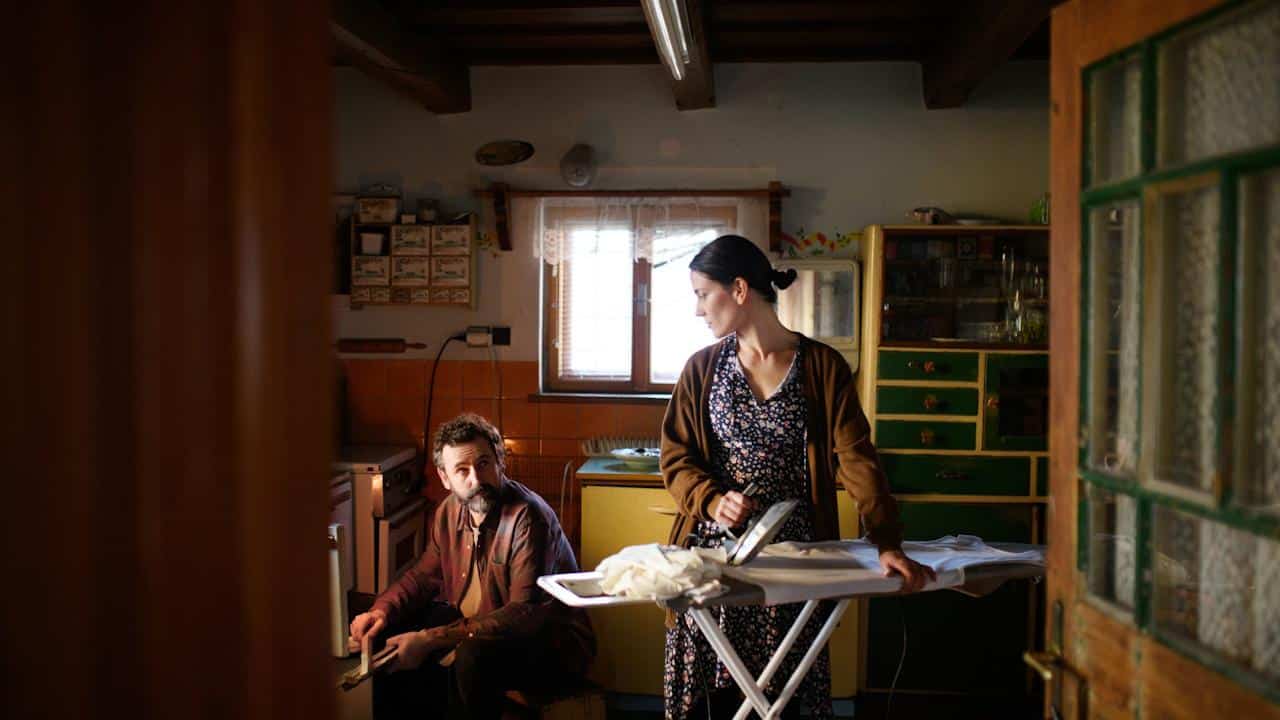 A woman is ironing in a rustic kitchen while a man sitting nearby looks towards her.