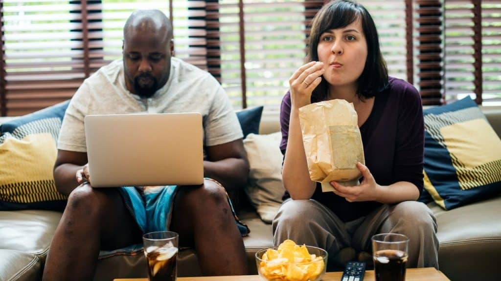 A man uses a laptop while a woman eats from a paper bag on a couch.
