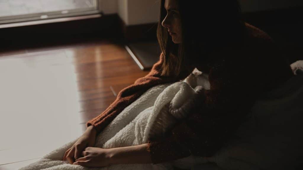 A person sitting on the floor wrapped in a blanket, looking toward a window in soft light.