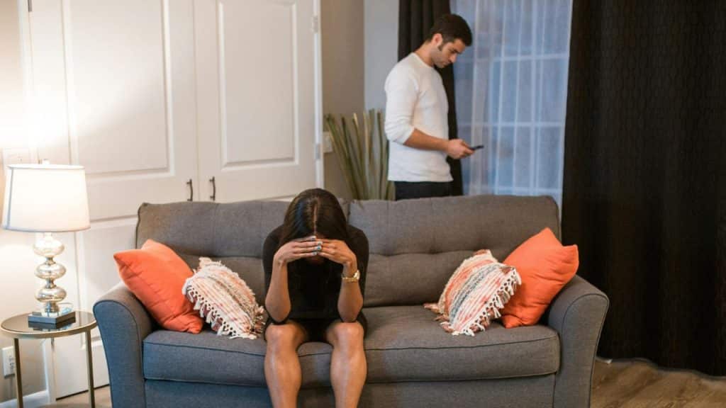 A woman sits on a couch covering her face while a man stands behind her using a phone.