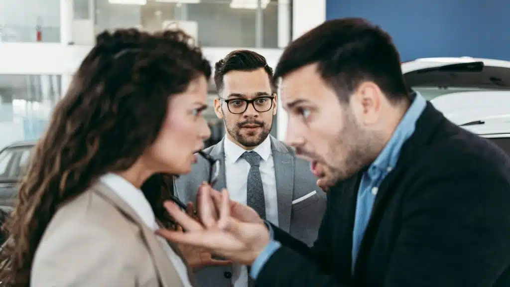 Two people speak intensely to each other while a man in a grey suit looks on.