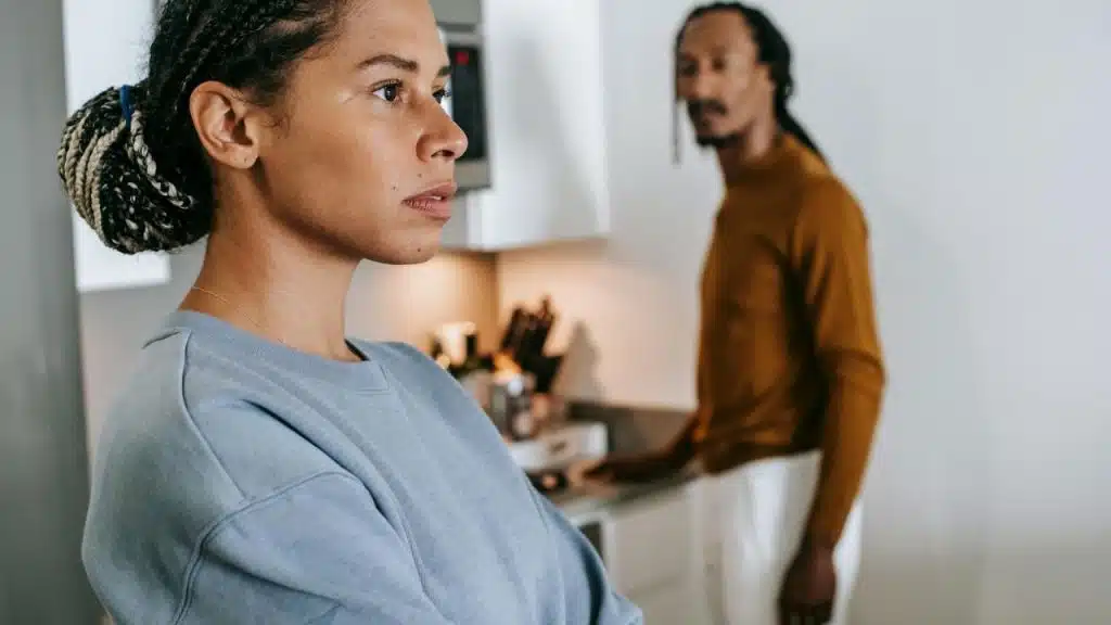 A woman with braided hair looks away while a man stands blurred in the background kitchen.