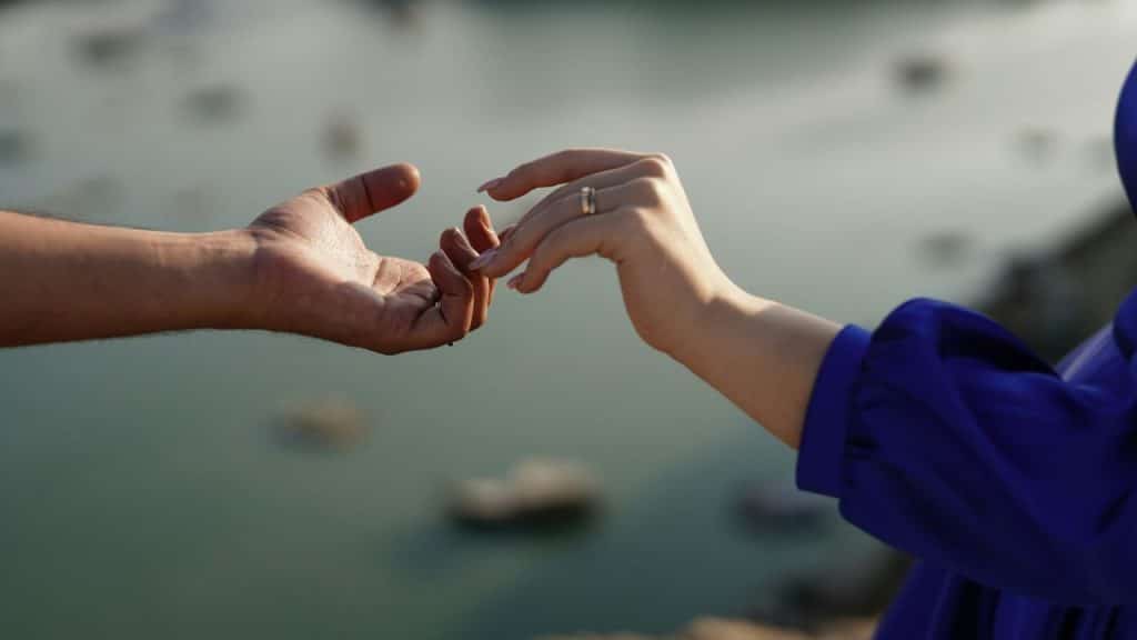 A person's hand reaches out toward another person's fingertips over a blurred water background.