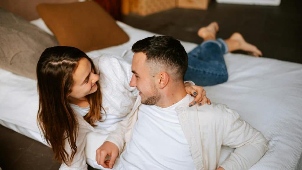 A couple lying on a bed facing each other and smiling in a bright, warmly-lit bedroom.