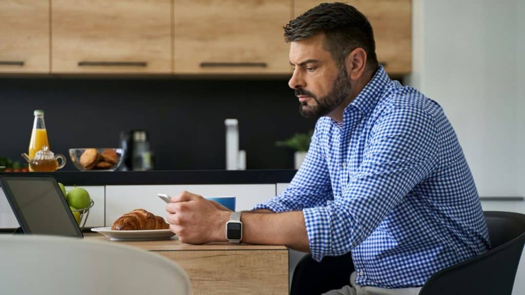 A man in a blue checkered shirt sits at a table looking at a smartphone.