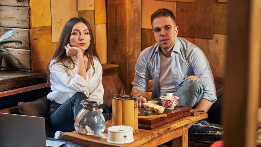 A man and woman sit at a low table with tea, looking forward with somber expressions.
