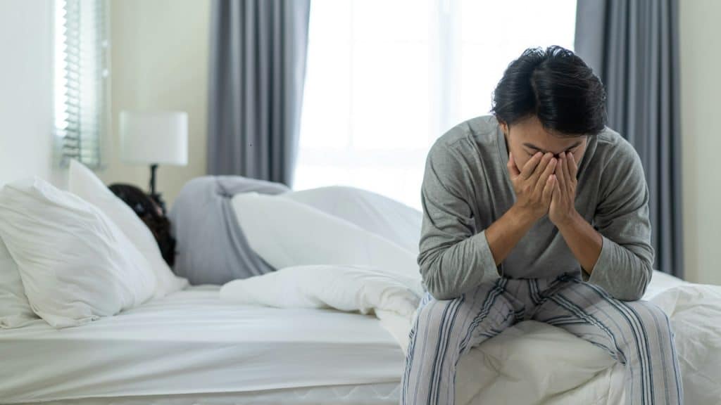 A frustrated man is sitting on one side of the bed while his wife is lying on the other side with her face on the opposite side.