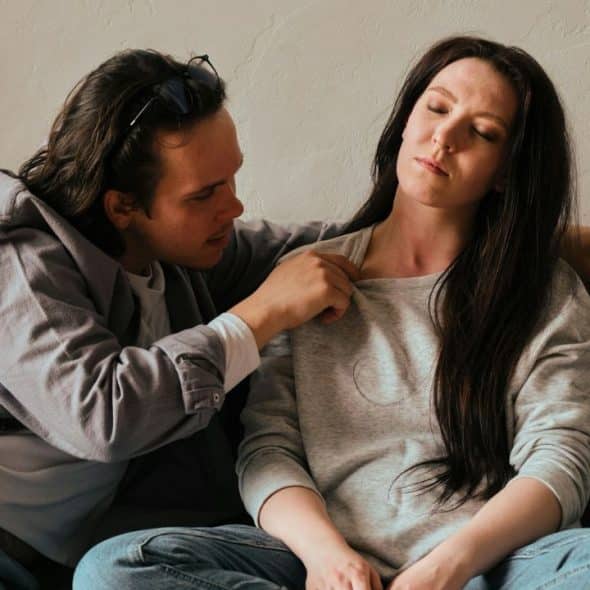 A man sitting on a sofa reaches out to pull the collar of a woman's sweatshirt.