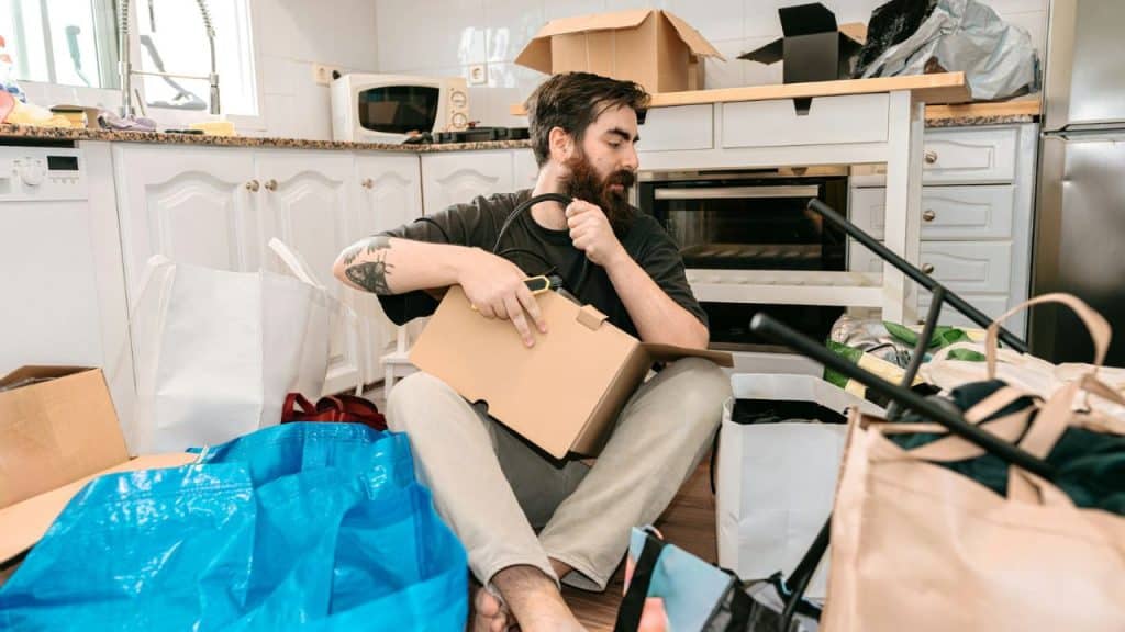A bearded man sits on a kitchen floor surrounded by open boxes and bags.