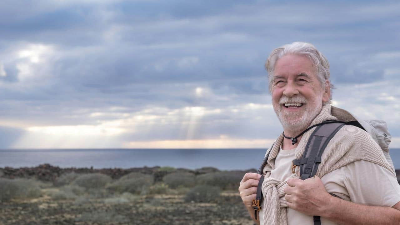 An elderly man with a backpack smiling by a rocky shoreline.
