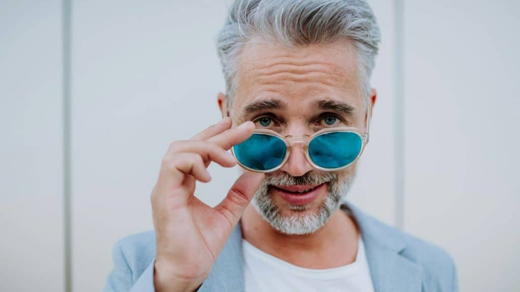 A man with grey hair and a beard lowers his blue-tinted sunglasses to look forward.