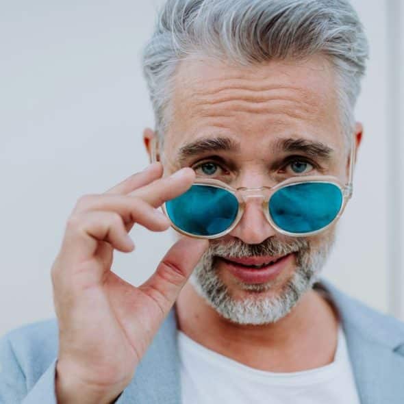 A man with grey hair and a beard lowers his blue-tinted sunglasses to look forward.