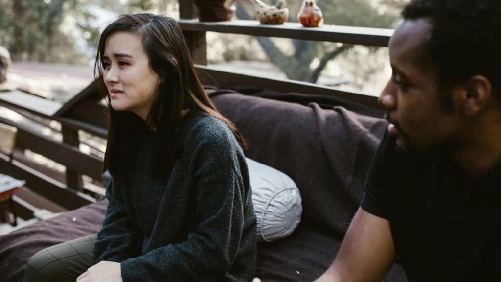 A woman with a sad expression sits on a porch while a man speaks nearby.