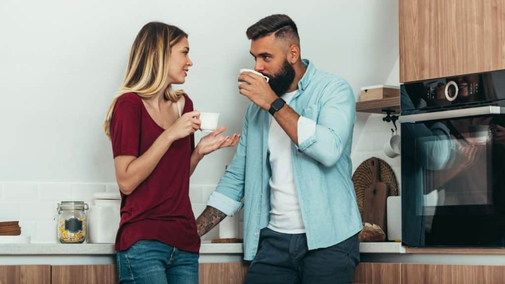 A woman in a red shirt talks to a bearded man while they both hold white mugs.