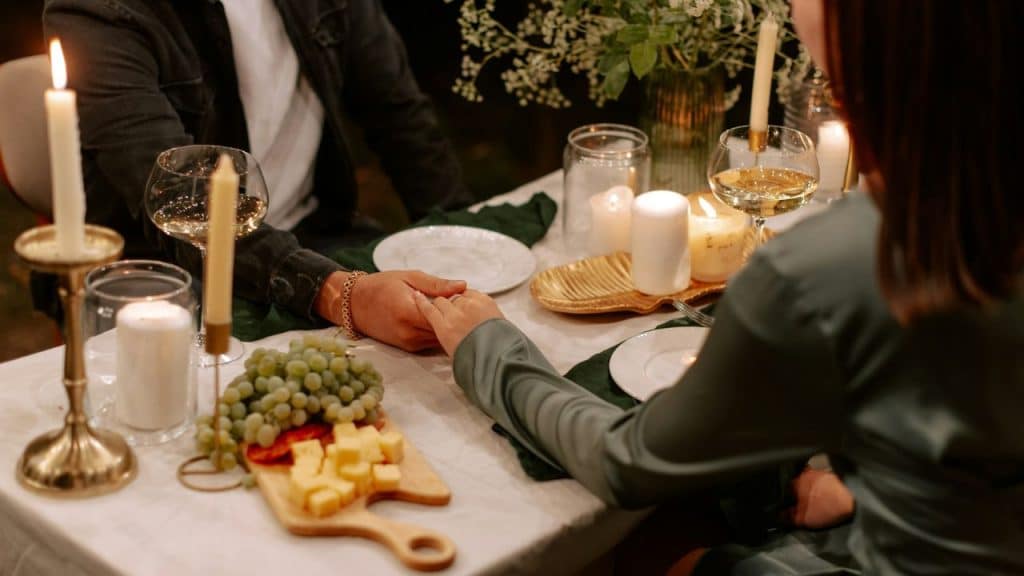 Two people hold hands across a candlelit dinner table with wine, cheese, and grapes.