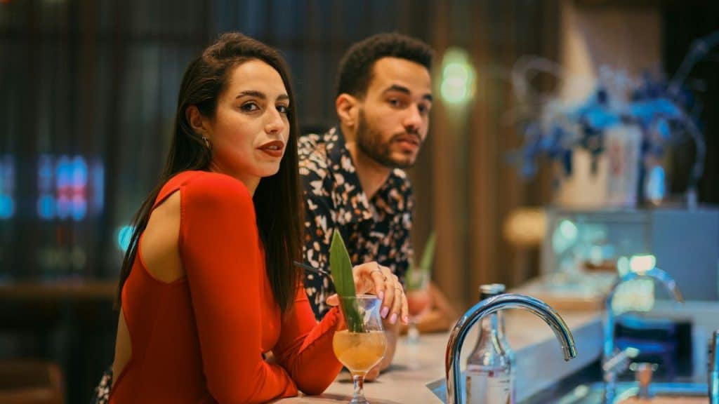 A man and woman sit at a bar counter with cocktails in a room.