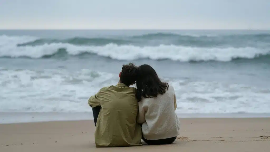 A couple sitting close and touching their heads while sitting on a beach by the sea.