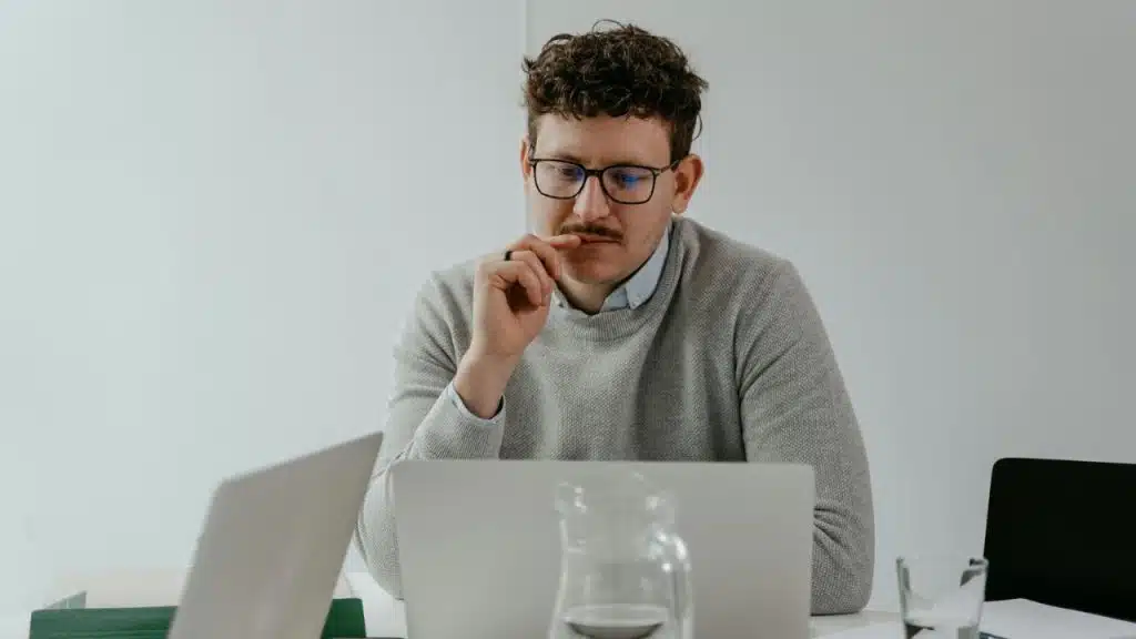 A man wearing glasses and a grey sweater sits at a table looking at a laptop.