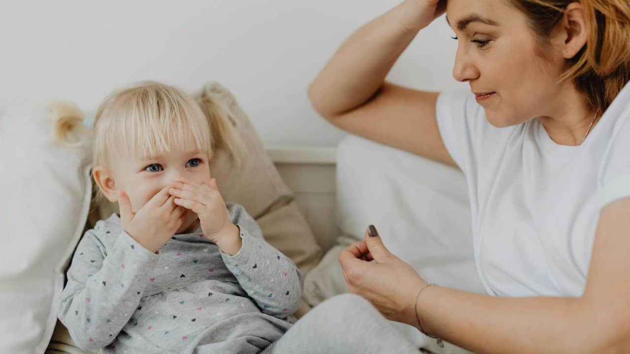 A woman looks toward a small child who is covering their mouth with both hands.