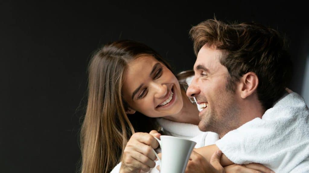 A smiling couple in white robes sharing a warm moment with a mug.