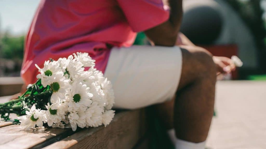 A bouquet of white daisies rests on a wooden bench next to a person sitting down.