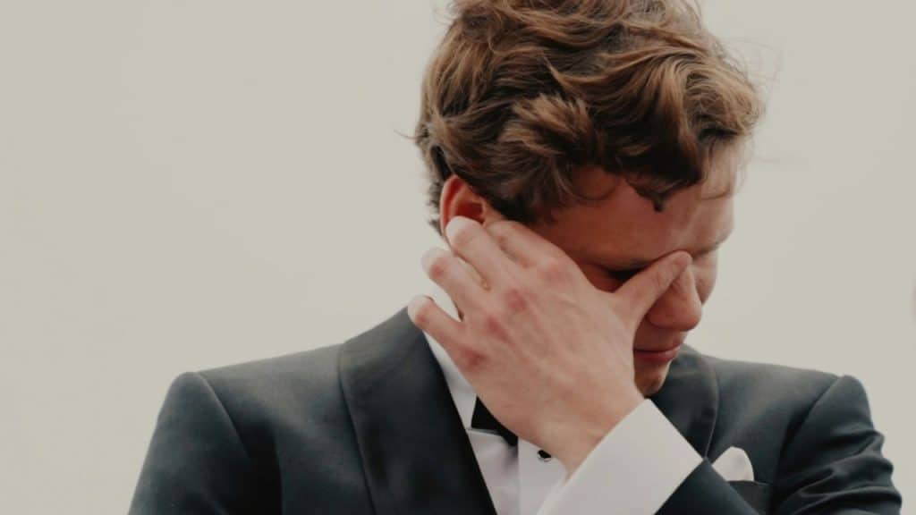A man in a tuxedo covers his eyes with his hand against a plain background.