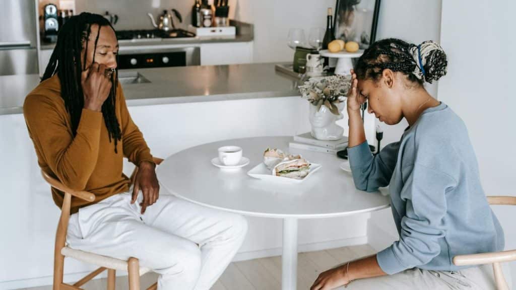 A man and woman sit at a white table with food, both looking down and away.