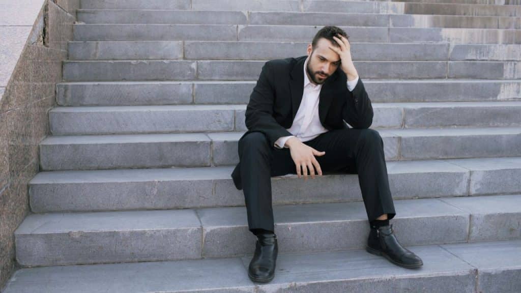 A man in a black suit sits on concrete stairs with one hand on his head.