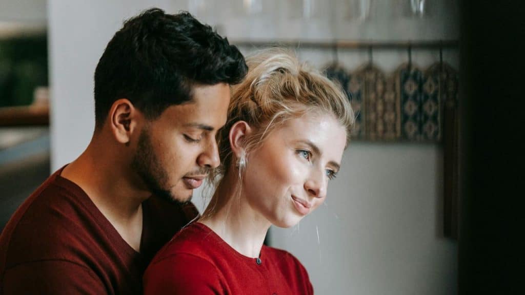 A man with dark hair leans close behind a blonde woman; both wear red shirts.