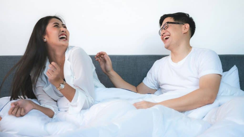 A man and woman sitting in bed together are laughing with their mouths wide open.