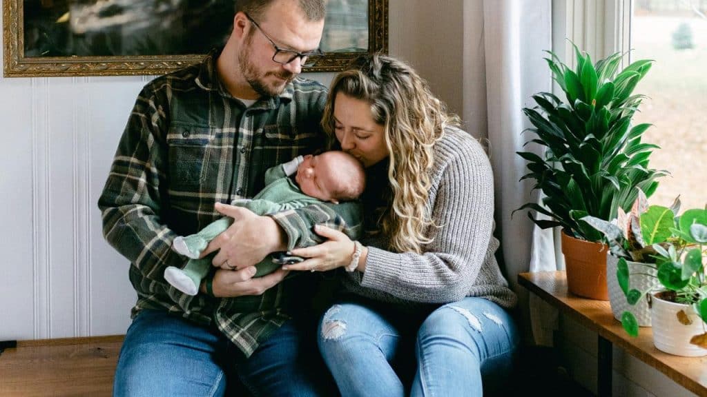 A couple sitting by a window cradling a newborn baby, with the mother kissing the baby’s head.
