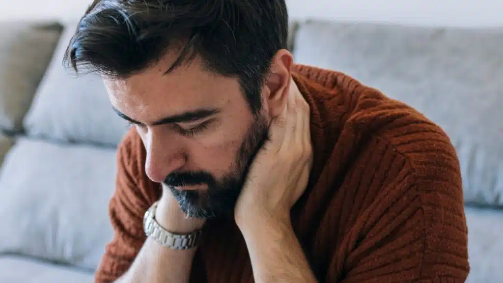 A man with a beard sits with his head bowed, holding the back of his neck.