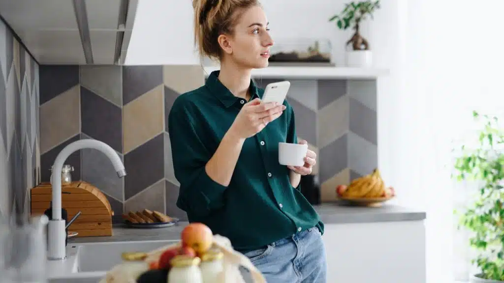 A woman in a kitchen holding a phone and a cup while looking to the side.