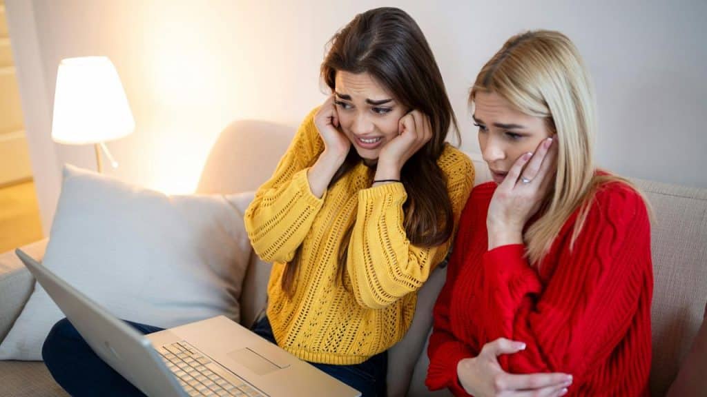 A pair of worried women sitting on a couch while looking at a laptop.
