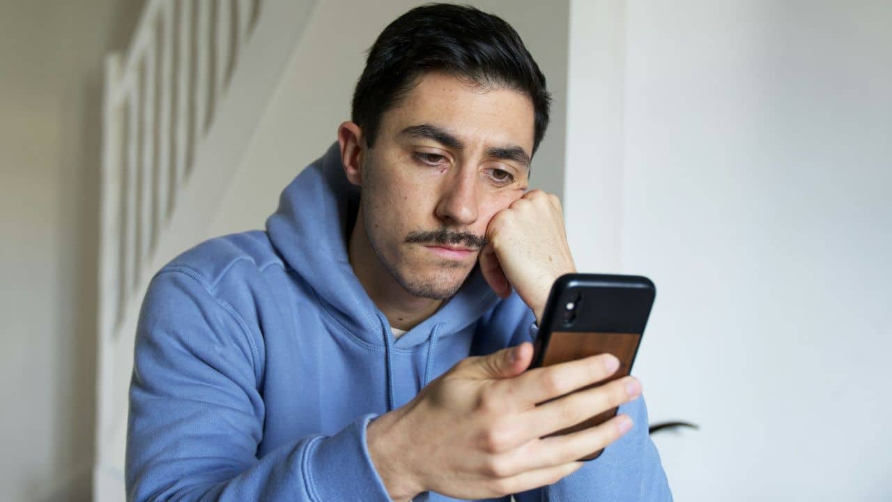 A man in a blue hoodie rests his head on his hand while looking at a smartphone.
