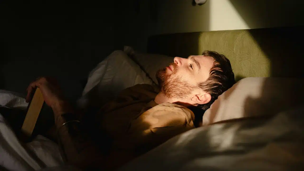 A bearded man lies in bed with a book, staring upward in a sunlit room.