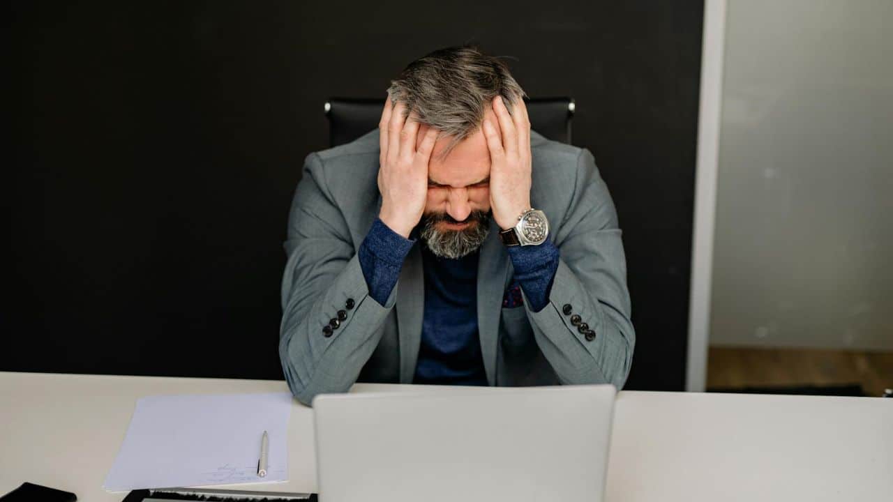 A bearded man in a grey suit sits at a desk holding his head in his hands.