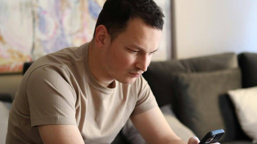 A man in a beige shirt sitting on a couch and looking intently at his phone.
