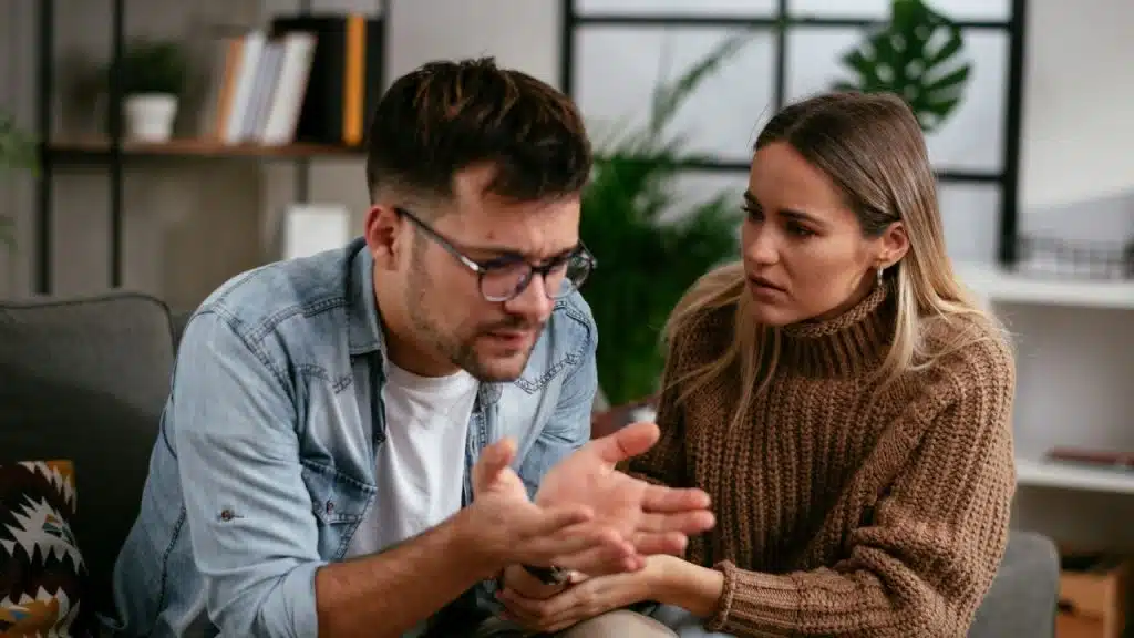 A man in a denim shirt gestures with his hands while a woman looks on with concern.
