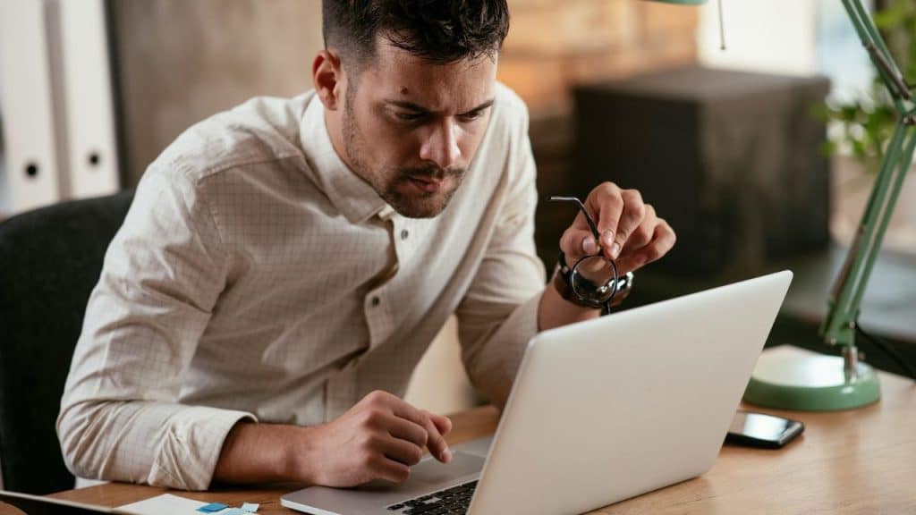 A man sitting at a desk holds glasses while looking intently at a laptop screen.