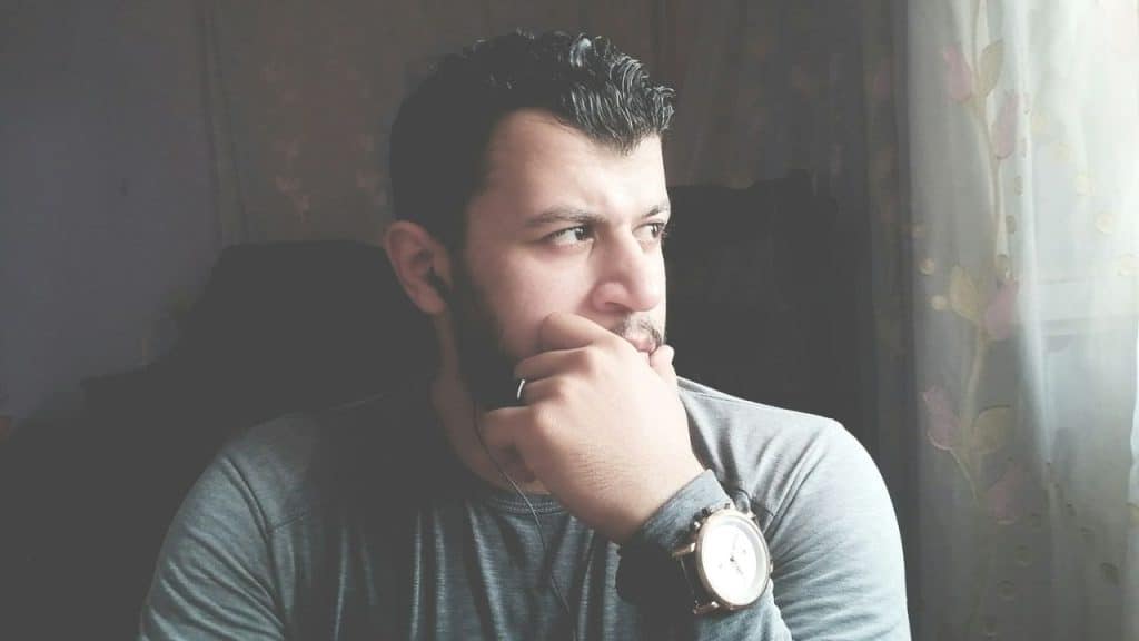 A man with a beard sits indoors, looking out a window with his hand on his chin.