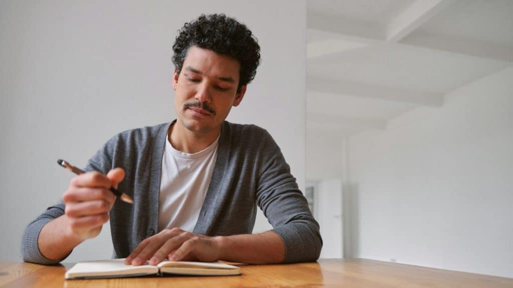 A man with curly hair and a mustache sits at a table writing in a notebook.